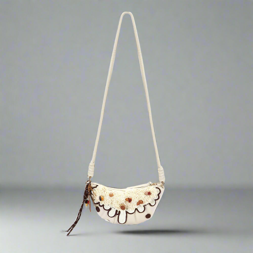 A cream-colored croissant-shaped shoulder bag with brown accents and a chain-ribbon tassel.