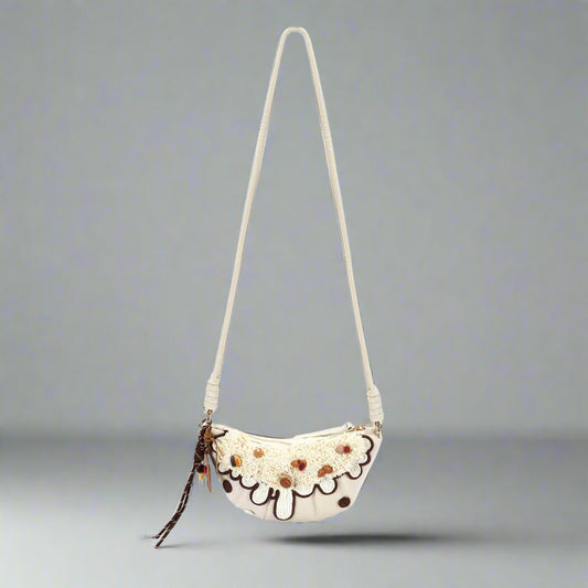 A cream-colored croissant-shaped shoulder bag with brown accents and a chain-ribbon tassel.