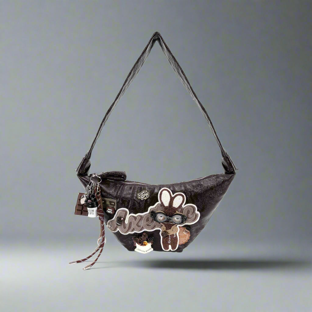 A crescent-shaped handbag with a dark brown textured finish, featuring a large plush bunny patch with 3D ears and silver glasses, and coffee-themed charms including a takeaway cup and chocolate bar.