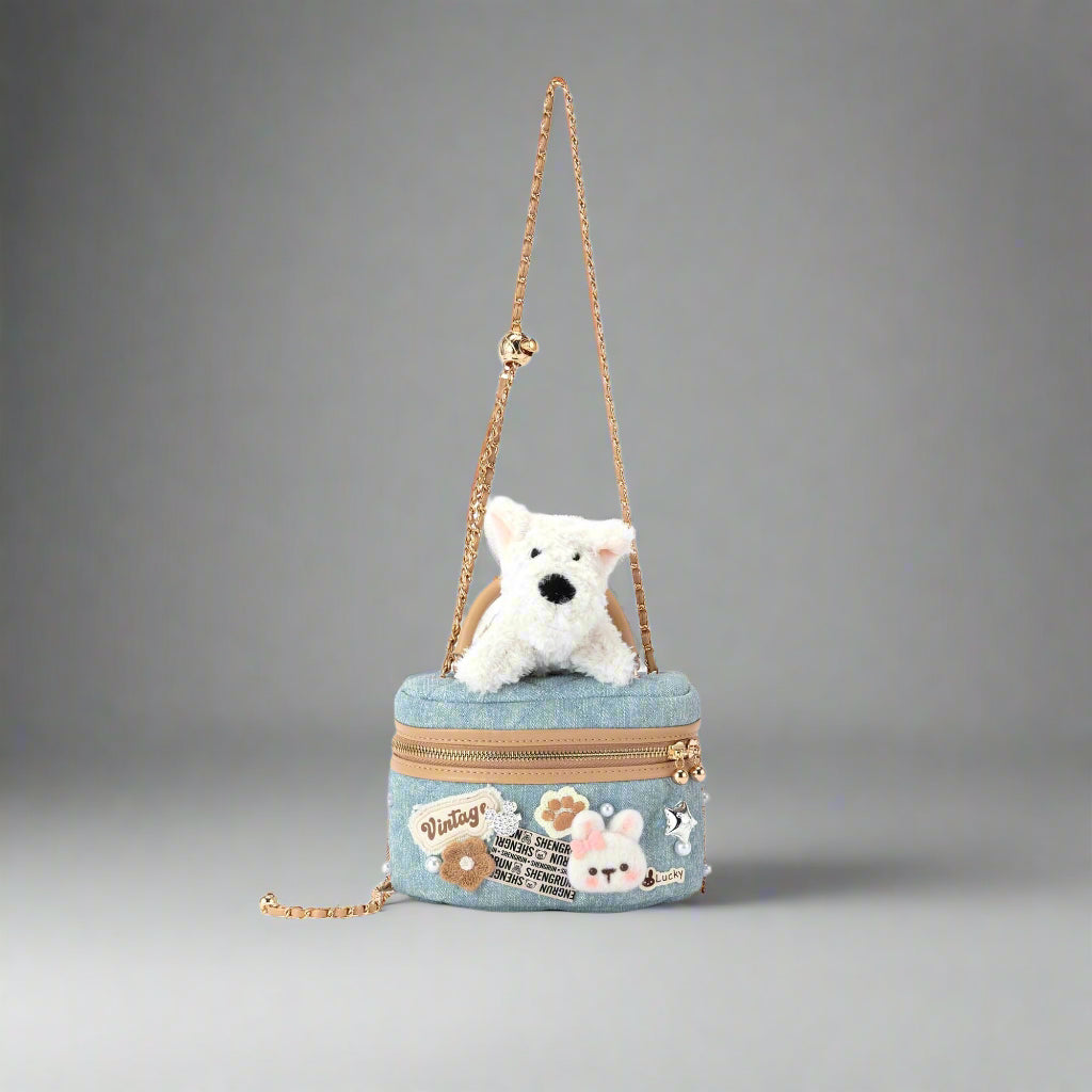 A crossbody cosmetic bag with a denim texture, featuring a plush terrier dog detail, and embellished with assorted 3D appliques and faux pearls. It has an adjustable gold-tone chain strap and a compact rectangular silhouette.
