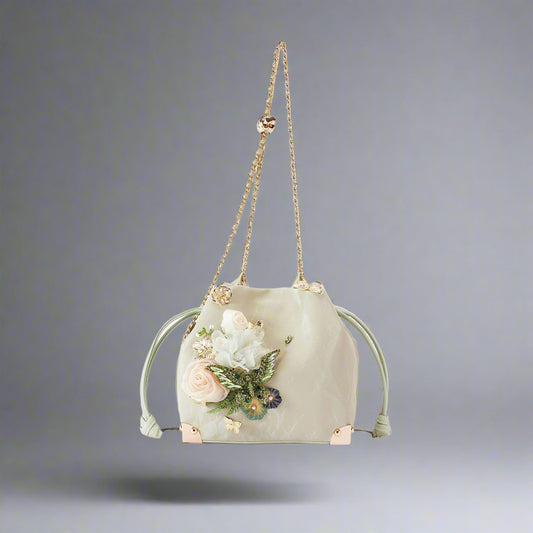 A sage green bucket bag with satin-textured body, 3D floral applique embellishments, butterfly brooches, and golden corner feet. It features an adjustable chain strap and a convertible design.