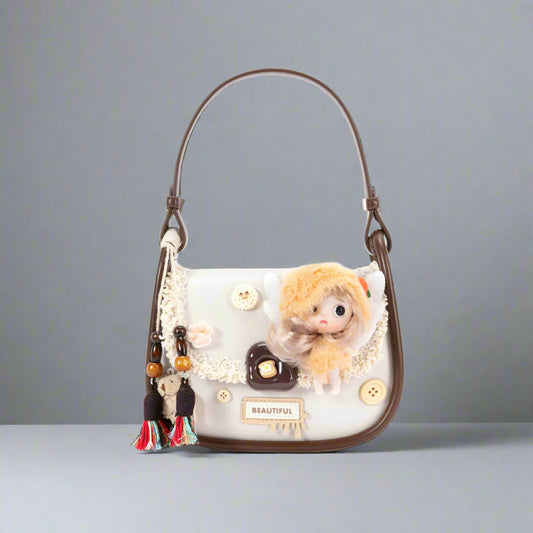 A vintage-fantasy style saddle bag with a plush doll embellishment, featuring a golden-yellow hood, white angel wings, and mixed button accents. The bag has a soft cream body with dark chocolate brown trim and an adjustable strap.
