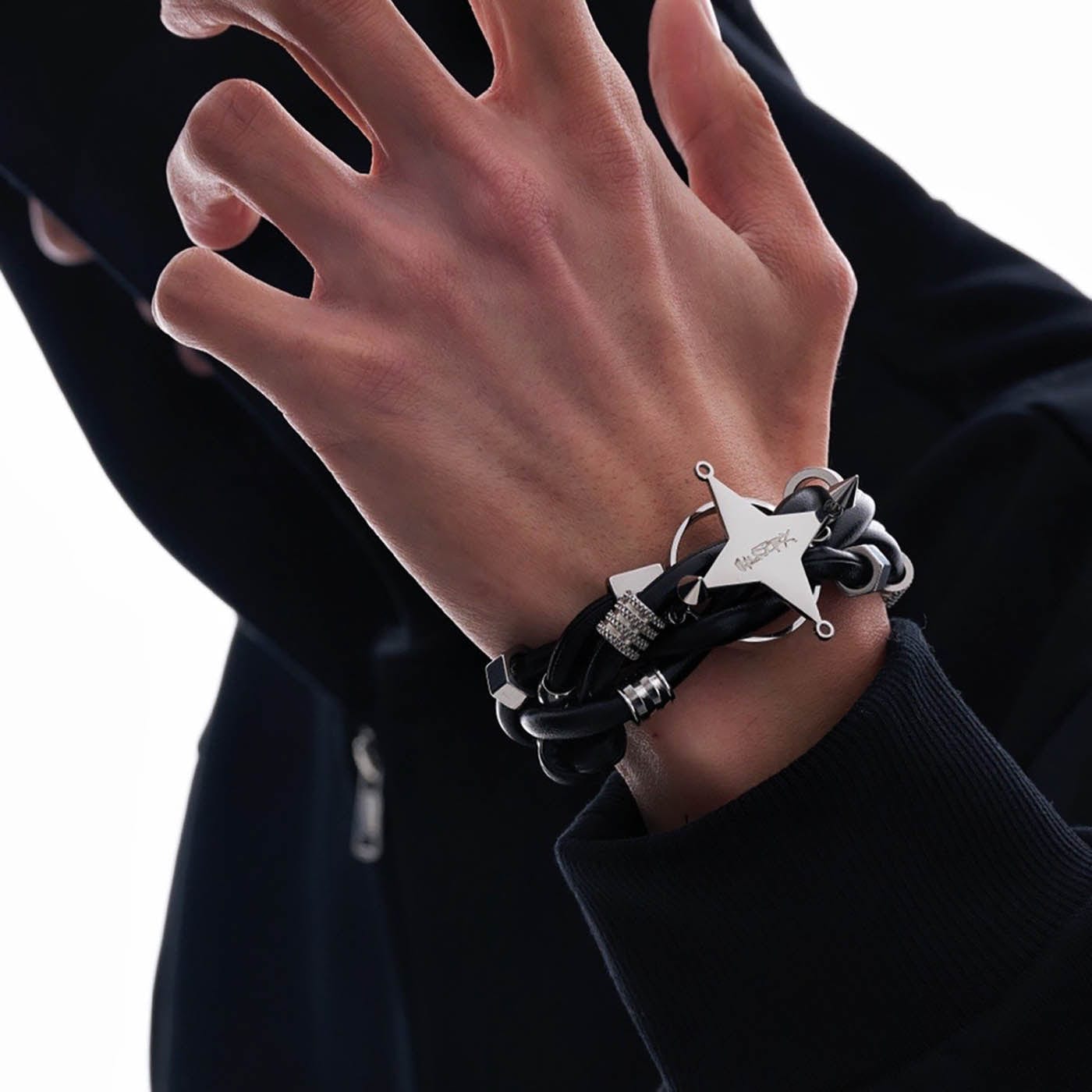 A person's wrist wearing a black braided faux leather bracelet with a silver four-point star centerpiece and metal block details.
