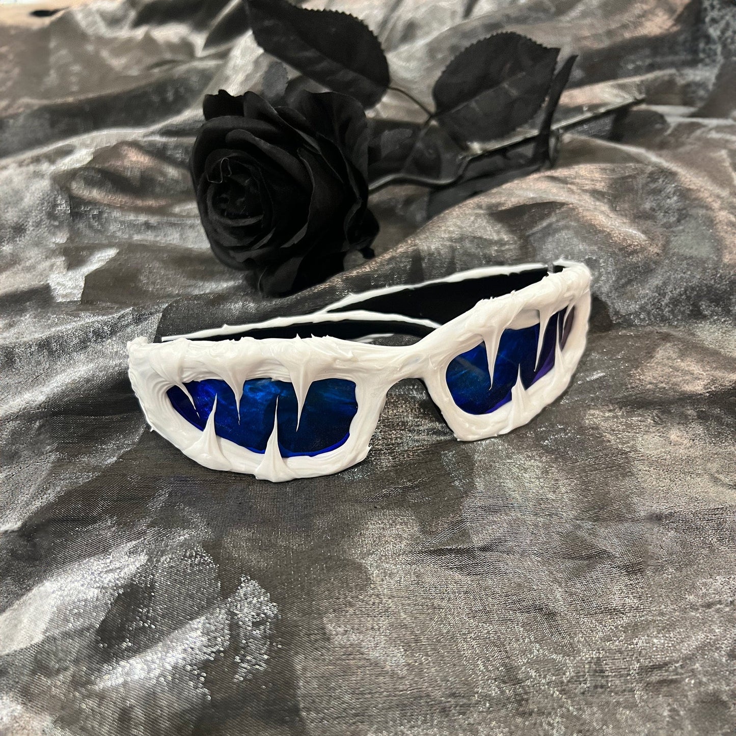 A pair of blue resin sunglasses with a unique design, placed on a surface with a black rose in the background.