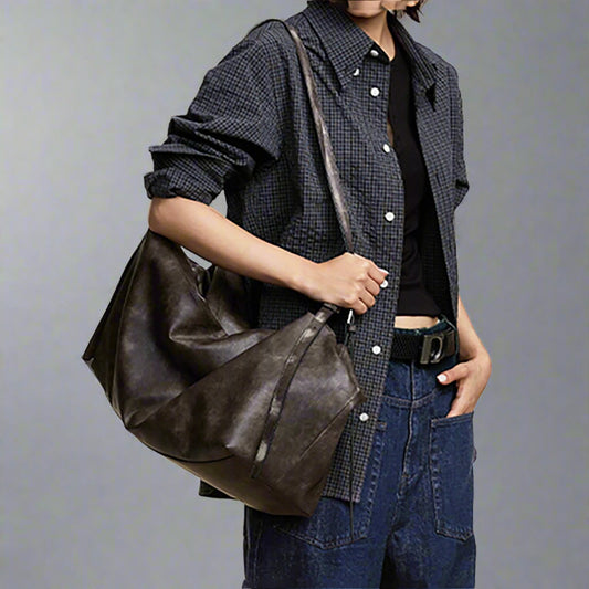 A person wearing a plaid shirt and blue jeans is holding a dark grey, double-strap rectangular PU shoulder bag.