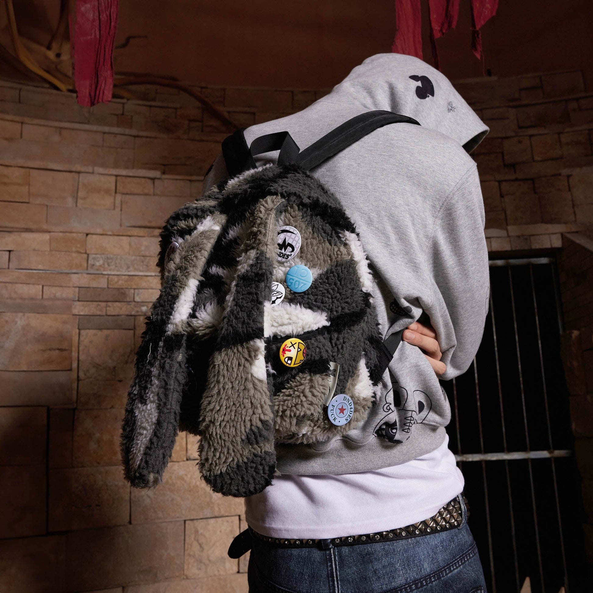 A person wearing a gray hoodie and white t-shirt is carrying a multicolored plush backpack with a camouflage design and stitched bunny ears on top.