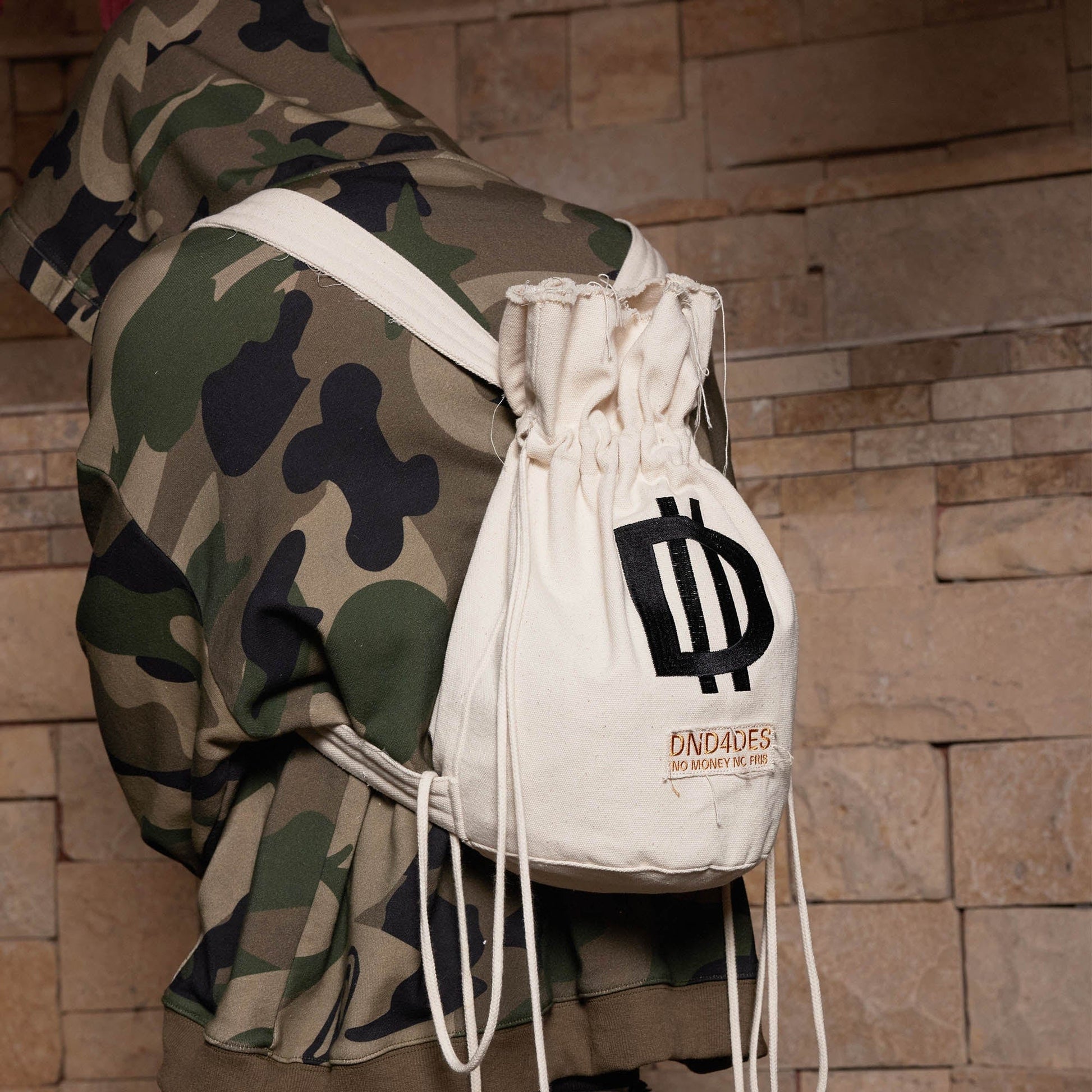 A person wearing a camouflage jacket is carrying a white drawstring bucket backpack with a logo on the front.